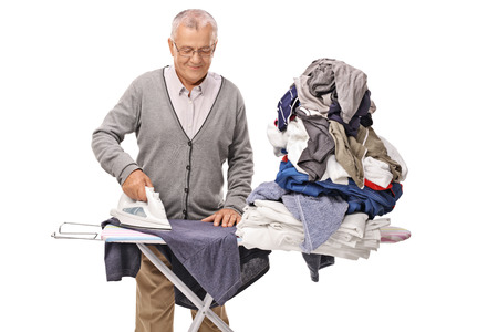 Cheerful senior man ironing a pile of clothes on an ironing board isolated on white backgroundの写真素材