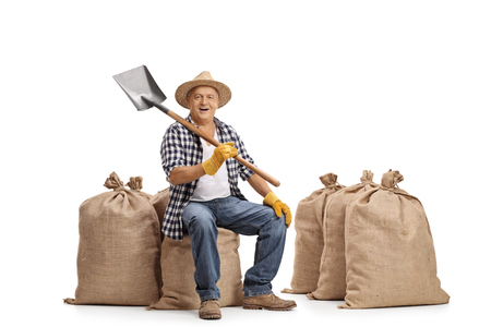 Mature farmer sitting on a burlap sack and holding a shovel isolated on white backgroundの写真素材