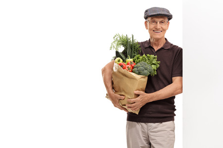 Elderly man holding a paper bag filled with groceries and leaning against a wall isolated on white backgroundの写真素材