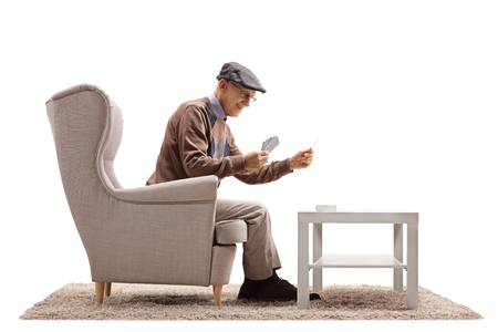 Senior seated in an armchair playing cards by himself isolated on white backgroundの写真素材