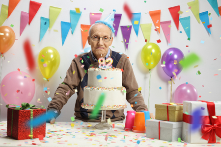 Sad elderly man with a birthday cake against a wall with decorationsの写真素材