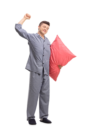 Full length portrait of a teenage boy in pajamas holding a pillow and stretching himself isolated on white backgroundの写真素材