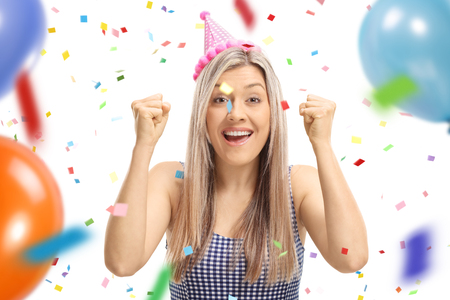 Young woman wearing a party hat gesturing happiness with confetti streamers and balloons flying around her isolated on white backgroundの写真素材