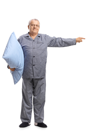 Full length portrait of an elderly man in pajamas holding a pillow and pointing isolated on white backgroundの写真素材