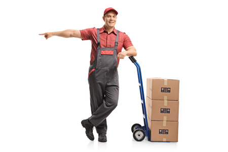 Full length portrait of a mover leaning on a hand truck with a stack of boxes and pointing isolated on white backgroundの写真素材