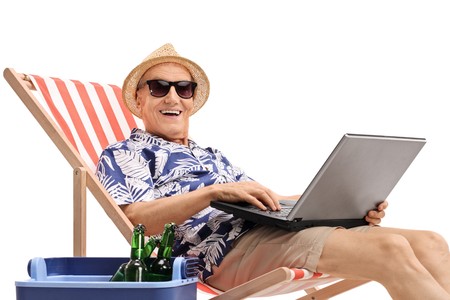 Old tourist with a laptop sitting in a deck chair next to a cooling box and looking at the camera isolated on white backgroundの写真素材