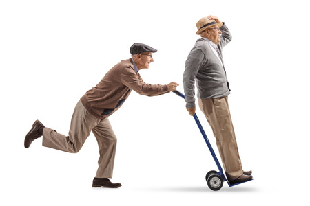 Full length profile shot of a senior pushing a hand truck with another senior riding on it isolated on white backgroundの写真素材