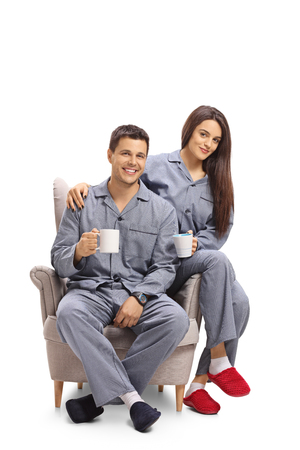 Young couple dressed in pajamas sitting in an armchair and holding cups isolated on white backgroundの写真素材