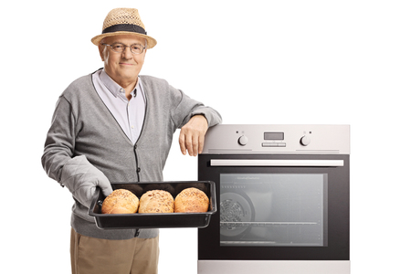 Mature man holding a tray with freshly baked bread loafs next to an oven isolated on whiteの写真素材
