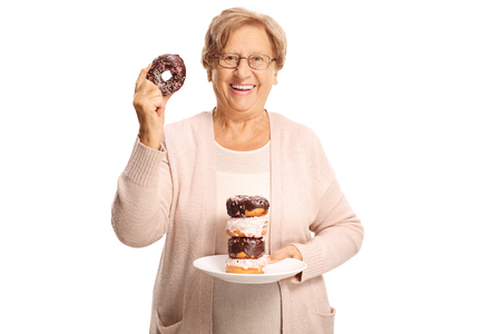 Cheerful elderly woman holding a plate of doughnuts in one hand and one doughnut in the other hand isolated on white backgroundの写真素材