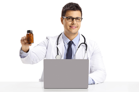 Young male doctor sitting with a laptop and holding a bottle of pills isolated on white backgroundの写真素材