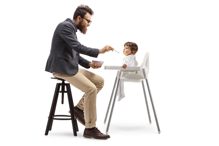 Full length profile shot of a father sitting and feeding his baby in a feeding chair isolated on white backgroundの写真素材