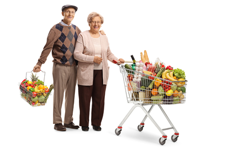 Full length portrait of a senior husband and wife posing with a shopping cart and a shopping basket with food isolated on white backgroundの写真素材