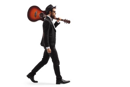Full length profile shot of a man in a suit walking with an acoustic guitar on his shoulder isolated on white backgroundの写真素材