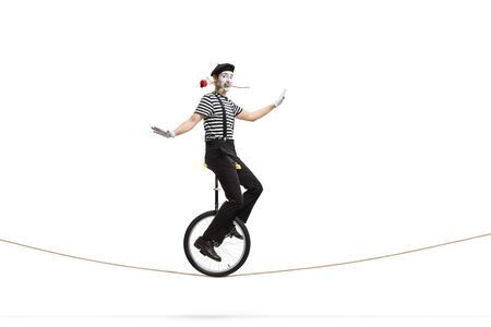 Full length shot of a mime riding a unicycle on a rope and holding a rose in his mouth isolated on whiteの写真素材