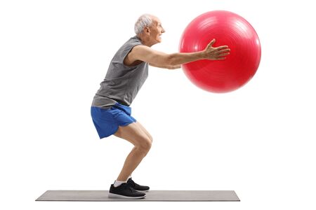 Full length profile shot of an elderly man exercising with a fitness ball on a mat isolated on white backgroundの写真素材