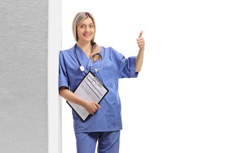 Young female doctor in a blue uniform leaning against wall and making a thumb up sign isolated on white backgroundの販売画像