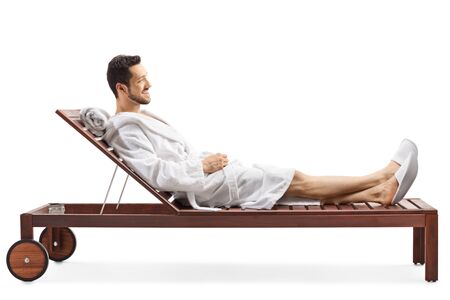 Full length profile shot of a young man in a bathrobe relaxing on a lounge chair isolated on white...