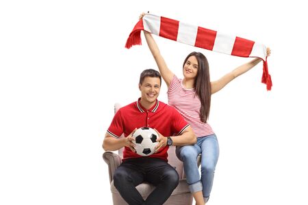 Young male and female soccer fans with a football and a scarf sitting in an armchair isolated on white backgroundの写真素材