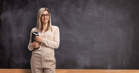 Young female teacher standing in front of a blackboard and holding booksの写真素材