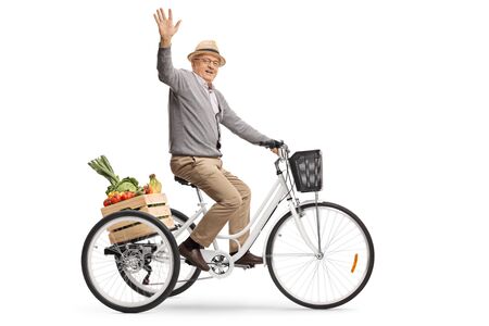 Elderly man riding a tricycle with a crate full of fruits and vegetables and waving at the camera isolated on white backgroundの写真素材