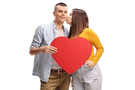 Young female kissing a guy holding a big red heart isolated on white background