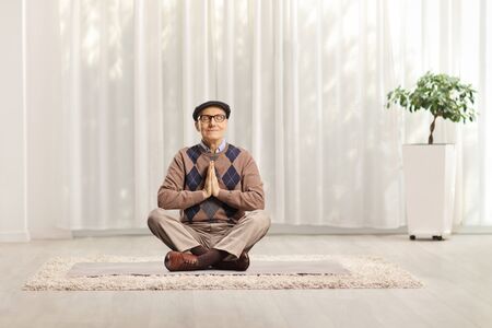 Senior man sitting on a mat with crossed legs meditating at homeの写真素材