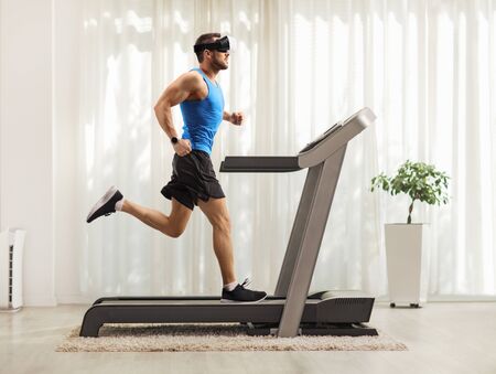 Full length profile shot of a young man running on a treadmill and wearing a vr headset at homeの写真素材