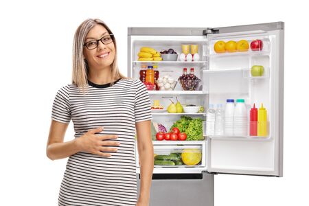 Young woman holding his hand on his stomach in front of a full refrigerator isolated on white backgroundの写真素材