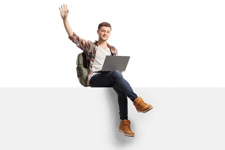 Male student with a laptop computer sitting on a blank panel and waving isolated on white backgroundの写真素材