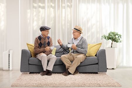 Two elderly men sitting on a sofa at home drinking tea and having a conversationの写真素材