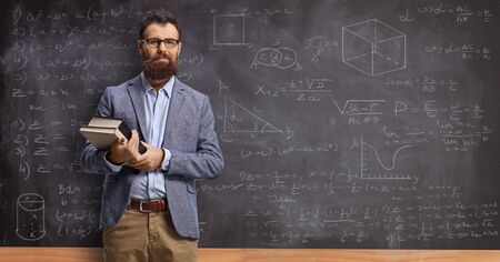 Male teacher holding books in front of a blackboard with math formulasの写真素材