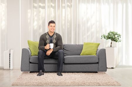 Full length portrait of a casual guy sitting on a sofa at home and holding a cup of teaの写真素材