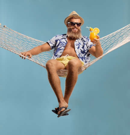 Bearded guy in shorts and shirt sitting on a hammock swing with a cocktail isolated on blue backgroundの写真素材