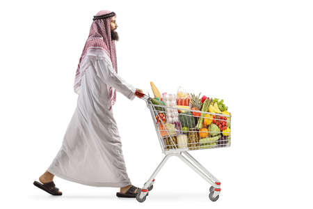 Full length profile shot of an arab man walking and pushing a shopping cart isolated on white backgroundの写真素材
