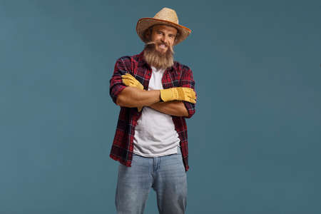 Bearded farmer with hat and gloves posing with crossed arms isolated on blue background