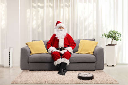 Full length portrait of santa claus at home sitting on a sofa and a robotic vacuum cleaner cleaning on the floorの写真素材