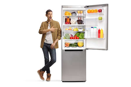 Full length portrait of a young man pointing at a fridge with food isolated on white backgroundの写真素材