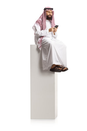 Arab man using a smartphone and sitting on high white column isolated on white backgroundの写真素材