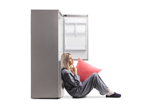Young woman in pajamas sitting on floor and leaning on an empty fridge isolated on white backgroundの写真素材