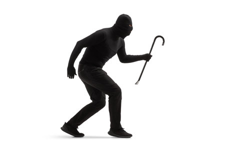 Full length shot of a burglar in black clothes with a balaclava and a crowbar walking slowly isolated on white backgroundの写真素材