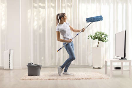 Young woman in front of a tv at home cleaning and dancing with a mop and playing air guitarの写真素材