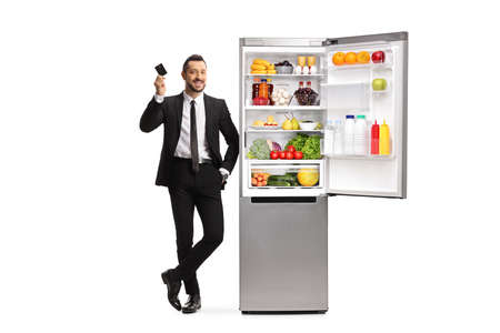 Full length portrait of a businessman holding a credit card and leaning on a fridge isolated on white backgroundの写真素材