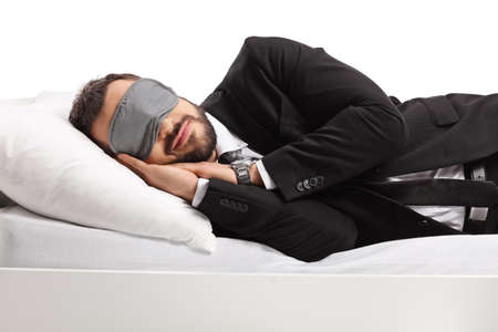 Businessman in a suit and tie sleeping on a bed and wearing a sleep mask isolated on white backgroundの写真素材