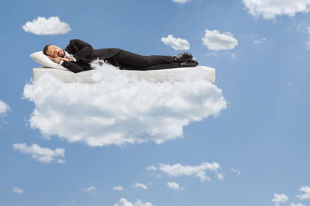 Businessman in a suit and tie sleeping on a mattress and floating on clouds and a blue skyの写真素材