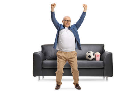 Mature football fan cheering in front of a black leather sofa isolated on white backgroundの写真素材