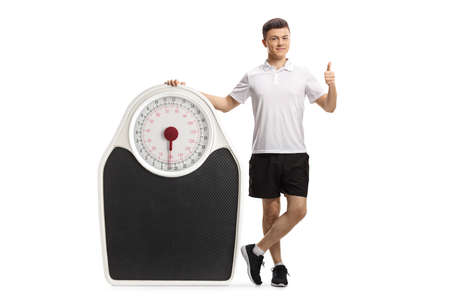 Full length portrait of a young man in sportswear leaning on a big scale and showing thumbs up isolated on white backgroundの写真素材