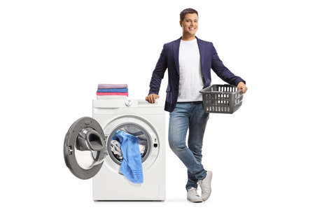 Young man with a laundry basket leaning on a washing machine with an open door isolated on white backgroundの写真素材