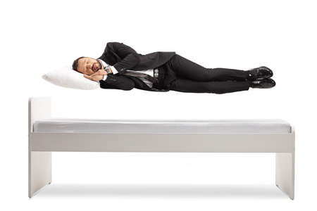 Man in a suit and tie sleeping and floating above his bed isolated on white background