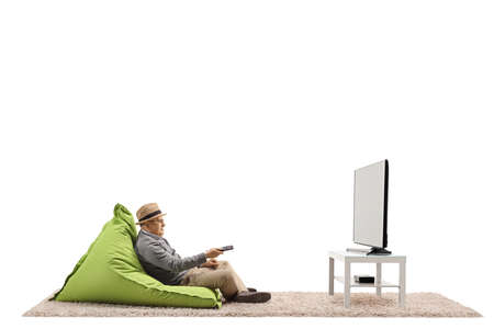 Full length shot of a senior gentleman with a remote controller sitting on a bean-bag chair and watching tv isolated on white backgroundの写真素材